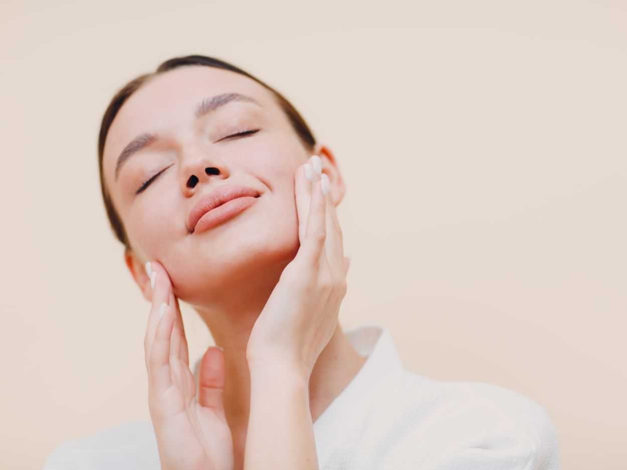 Woman touching her clear skin gently, eyes closed, on beige backdrop.